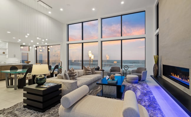 Modern open-plan living room with floor-to-ceiling windows overlooking ocean at sunset, gray sectional sofa, fireplace, and dining area.