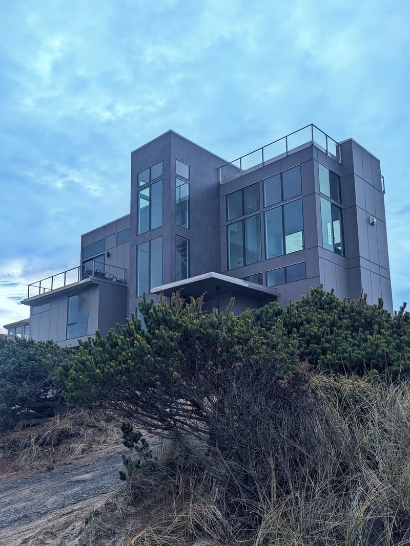Modern dark gray multi-story house with large windows and rooftop deck overlooking coastal dune vegetation.