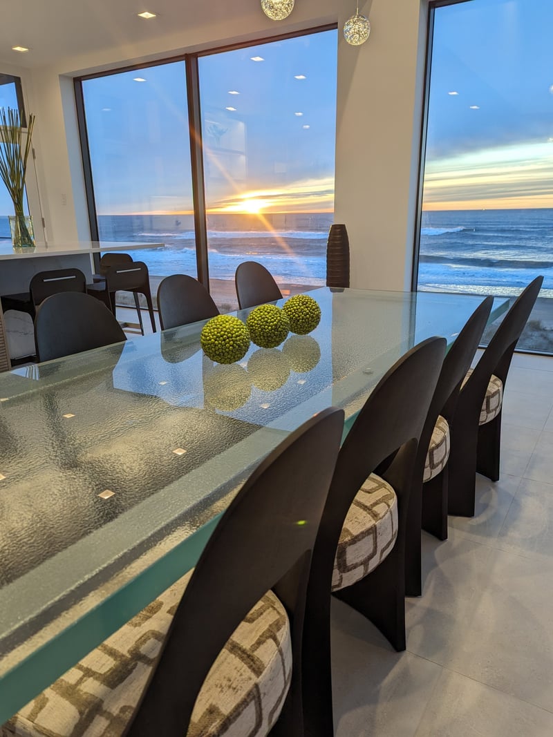 Modern dining room with glass table, black chairs, and floor-to-ceiling windows overlooking ocean sunset