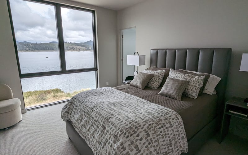 Modern bedroom with gray upholstered bed, lake and mountain views through large windows, bedside lamp, and cream armchair.