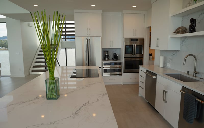 Modern kitchen with white cabinetry, marble countertops, stainless steel appliances, and island seating overlooking stairs and water view.