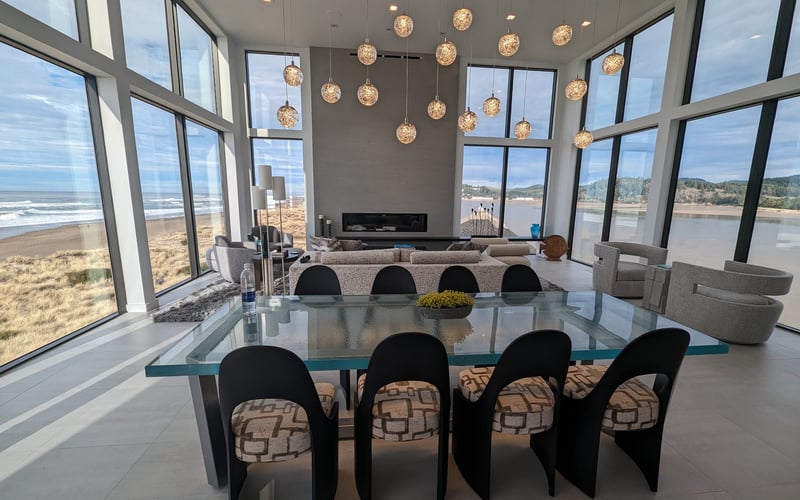 Modern open-plan living space with glass dining table, black chairs, gray sofa, hanging pendant lights, fireplace, and expansive windows overlooking beach and
