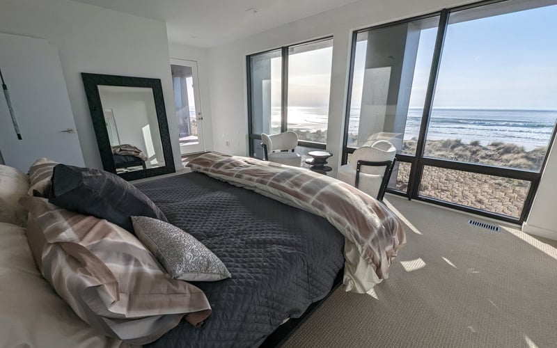 Modern bedroom with ocean and beach view through large windows, queen bed with dark bedding, and seating area