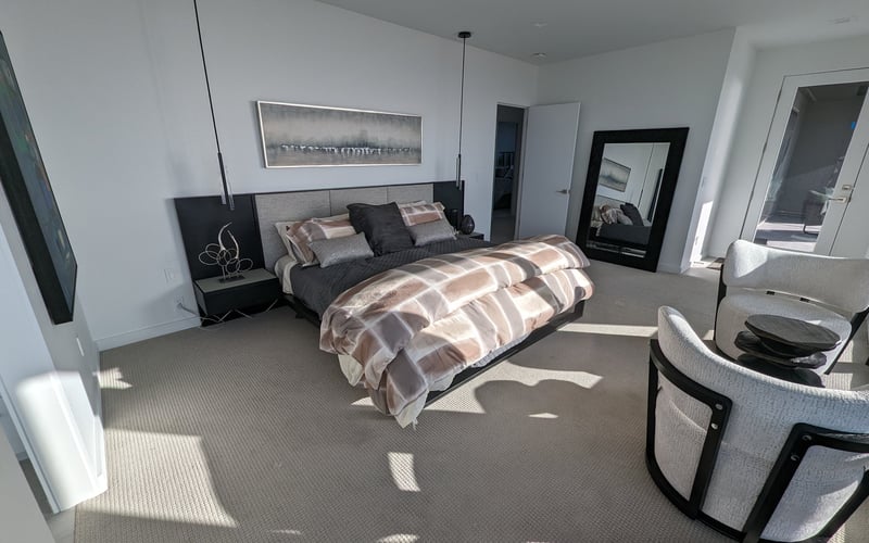 Modern master bedroom with striped bedding, black nightstands, pendant lights, framed artwork, and seating area with gray carpet flooring.