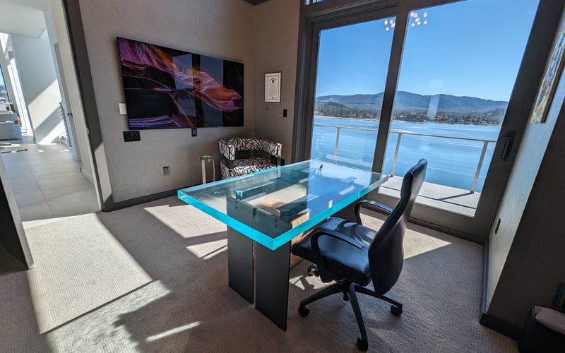 Modern office with turquoise glass desk, black chair, wall-mounted TV, and floor-to-ceiling windows overlooking a blue lake and mountains.
