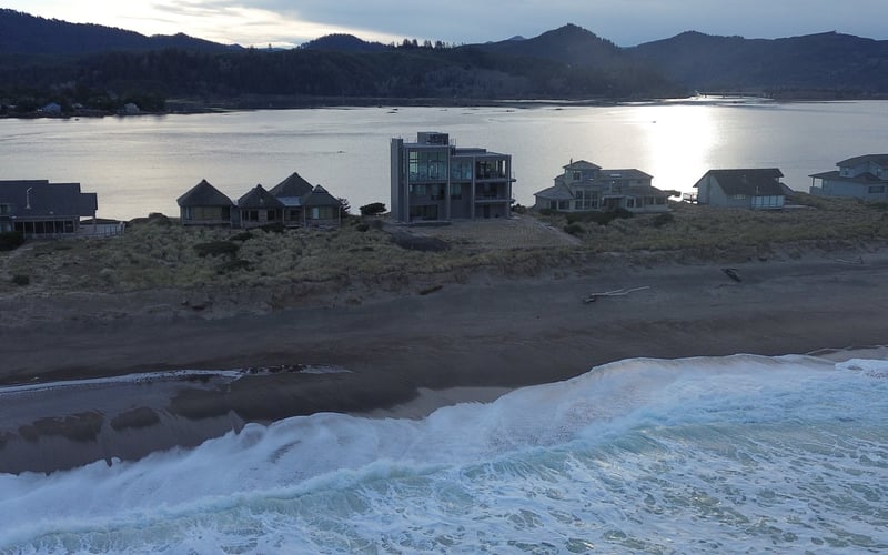Beachfront homes on sandy shore with waves in foreground, forested mountains and calm bay water beyond