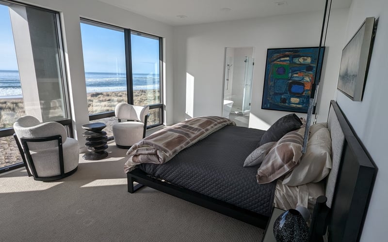 Modern bedroom with ocean and beach view through floor-to-ceiling windows, black bed, cream armchairs, and artwork on walls