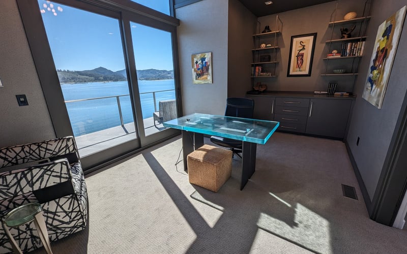 Modern office with glass desk, built-in shelving, and floor-to-ceiling windows overlooking blue water and mountains.