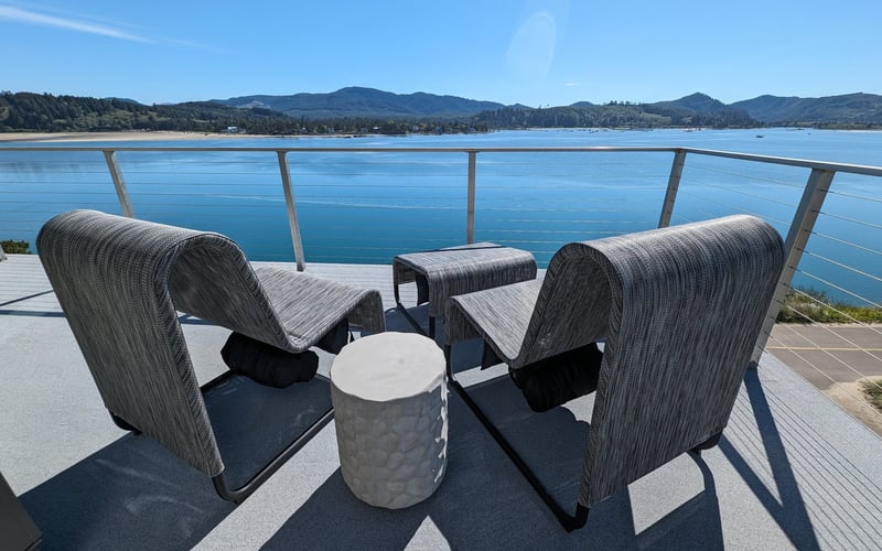 Waterfront deck with two woven lounge chairs, small table, and metal railing overlooking calm blue lake and forested mountains.
