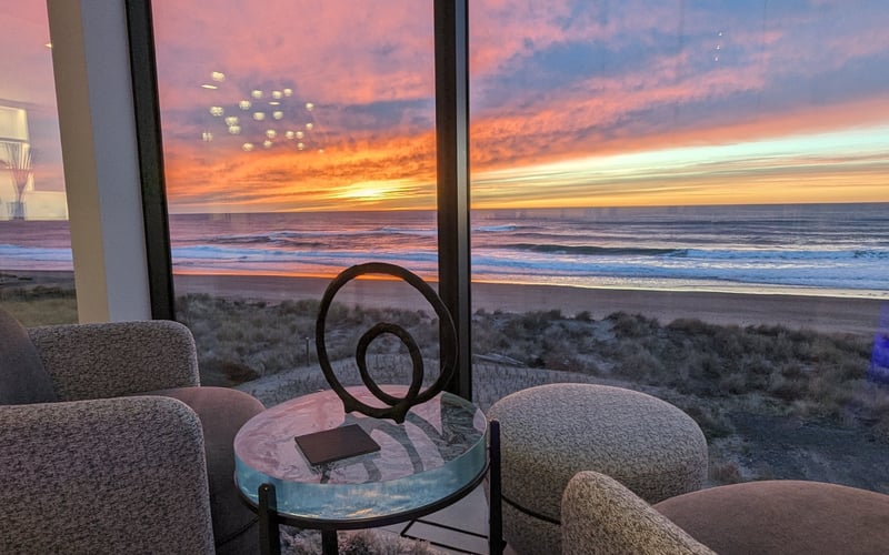 Modern lounge chairs and side table with ocean sunset view through floor-to-ceiling windows, beach and dunes visible below
