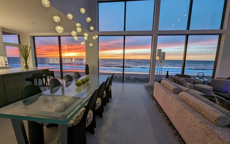Modern open-plan living room with dining area, beige sofa, and floor-to-ceiling windows overlooking ocean sunset at dusk