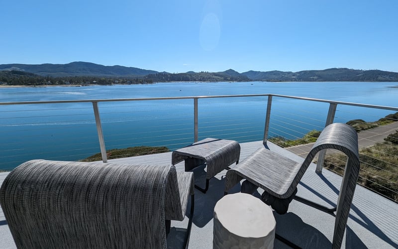 Waterfront deck with woven lounge chairs overlooking calm blue bay and forested mountains