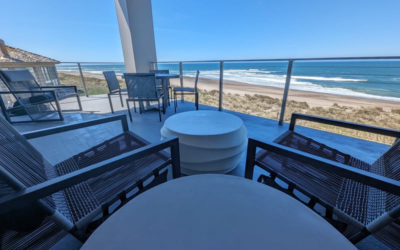 Oceanfront deck with round tables, lounge chairs, and glass railing overlooking sandy beach and blue ocean.